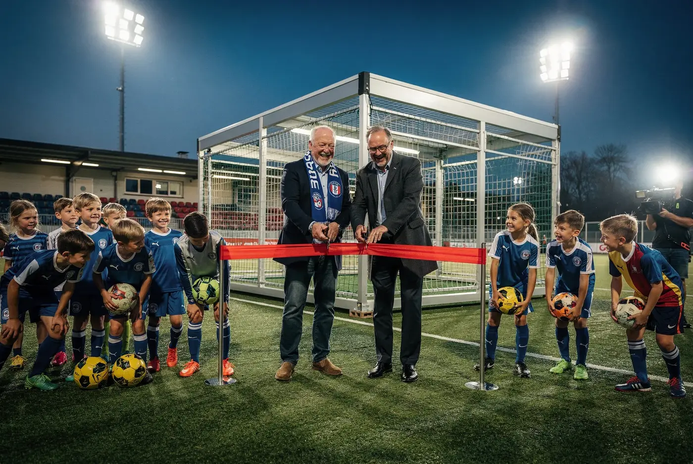 Vereinsvorstand und Unternehmer durchschneiden ein rotes Band vor der neuen Soccer-Arena, die Jugend wartet mit Bällen